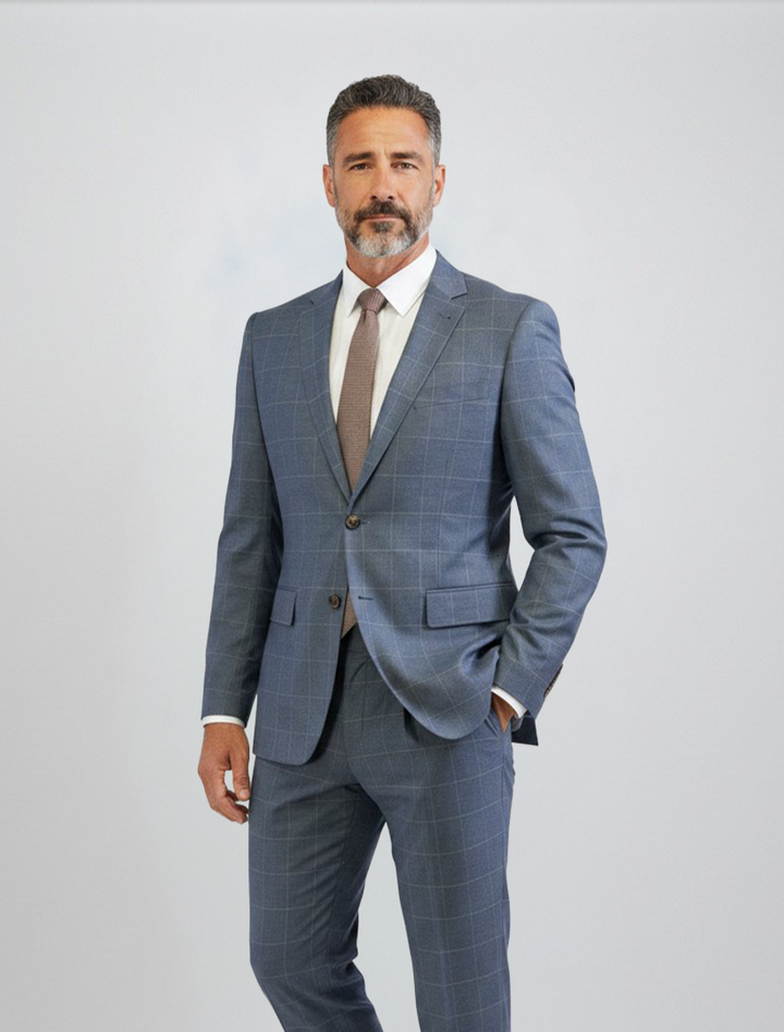 A man with short gray hair and a beard wears the FUBU Gravel Blue with Peach Windowpane 2-Piece Suit by Fubu The Colle, featuring a slim fit crafted from Tetron Rayon. Paired with a white shirt and brown tie, he stands against a plain light background with one hand in his pocket, while the suit’s stretch armhole provides both comfort and style.
