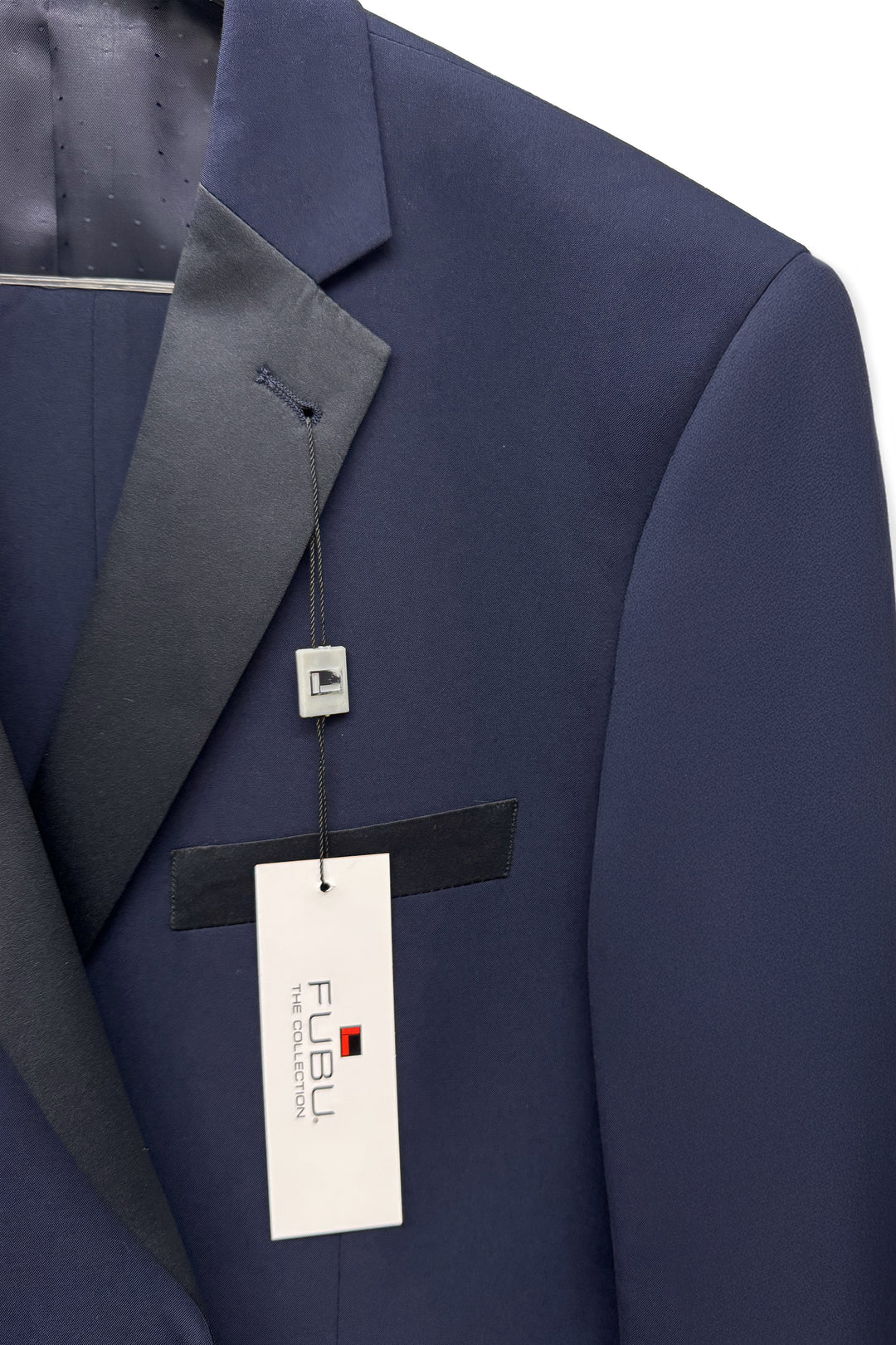 Close-up of the FUBU Classic Notch Lapel Navy Slim-Fit Tuxedo by Fubu The Colle, featuring black satin notch lapels, a hanging brand tag, and a string attached to the buttonhole.