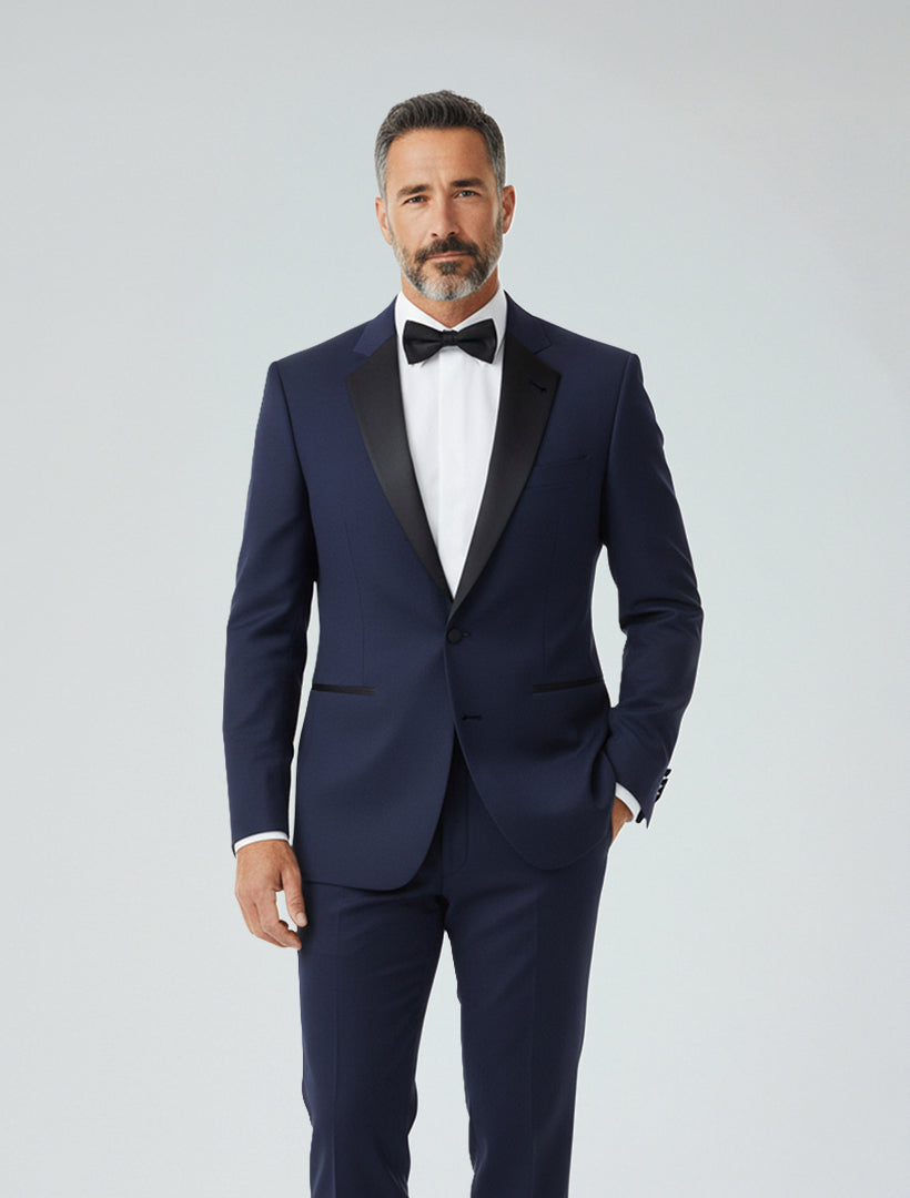 A man in a FUBU Classic Notch Lapel Navy Slim-Fit Tuxedo by Fubu The Colle, paired with a white dress shirt and black bow tie, stands against a plain light background.