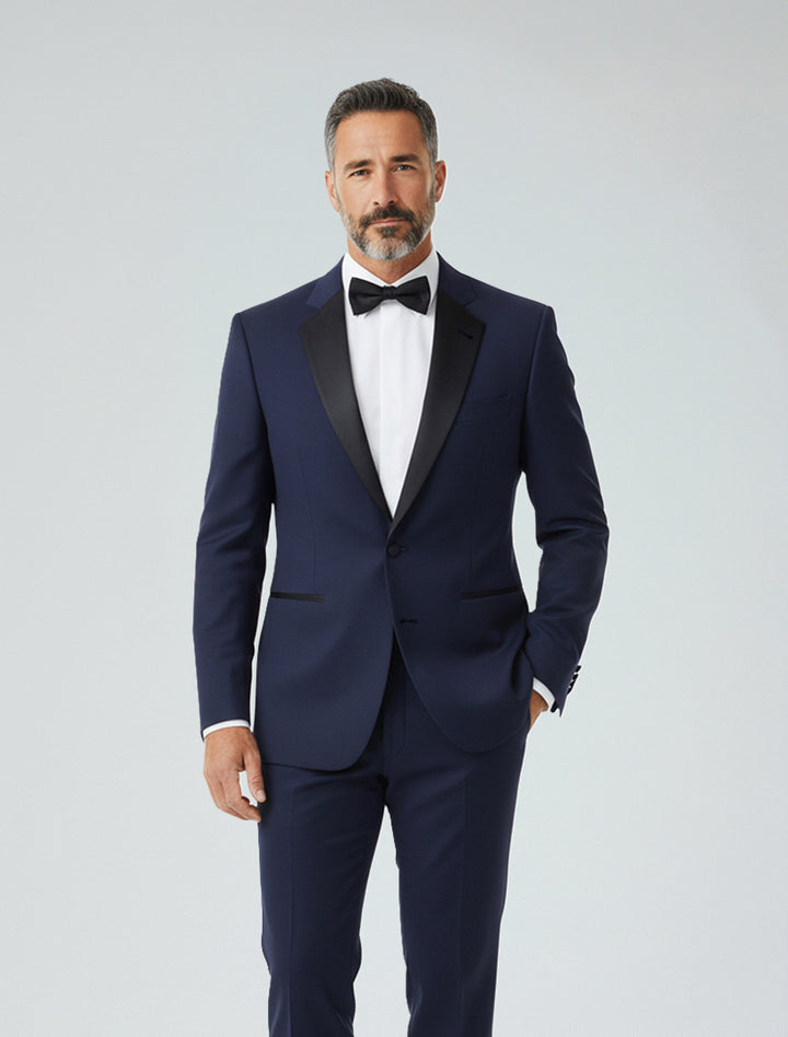 A man in a FUBU Classic Notch Lapel Navy Slim-Fit Tuxedo by Fubu The Colle, paired with a white dress shirt and black bow tie, stands against a plain light background.