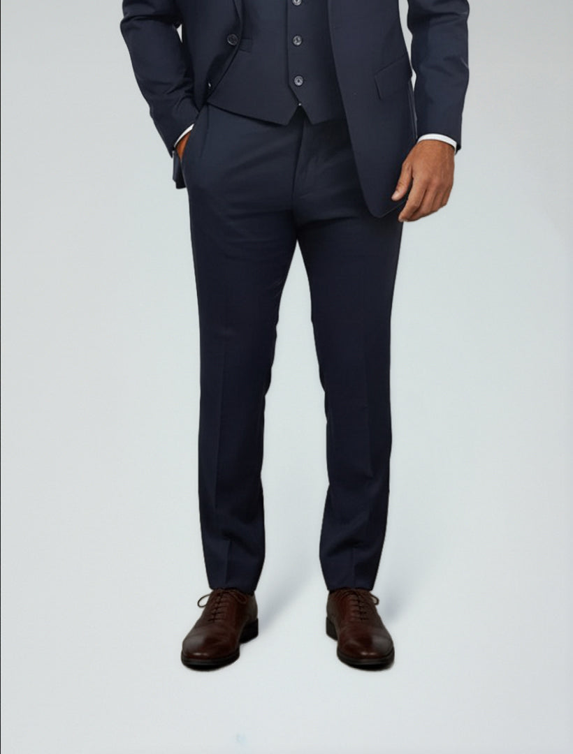 A man in Karako Separate Navy Men's Slim-Fit Suit Separates Pants, paired with a vest, white shirt, and brown dress shoes, stands with one hand in his pocket against a plain light background.