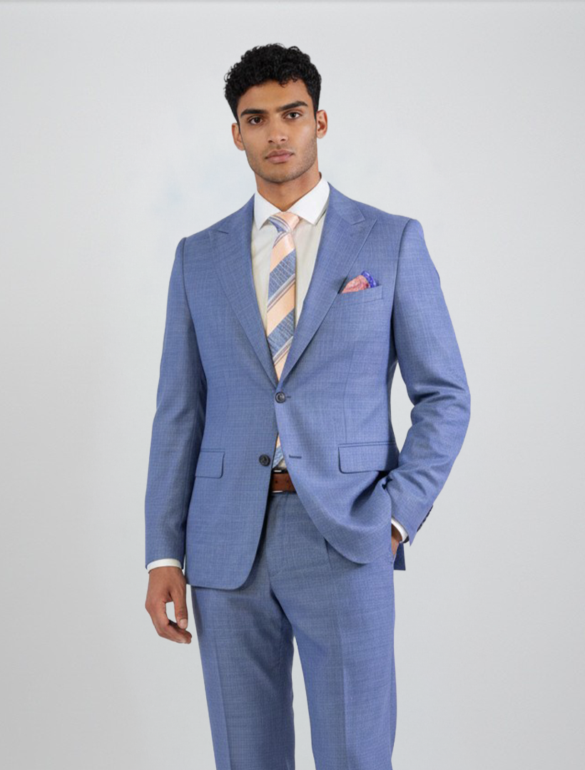 A man stands against a plain background wearing the FUBU Stone Blue Peak Lapel 2-Piece Suit by Fubu The Colle, styled with a white dress shirt, striped tie, and pink pocket square.