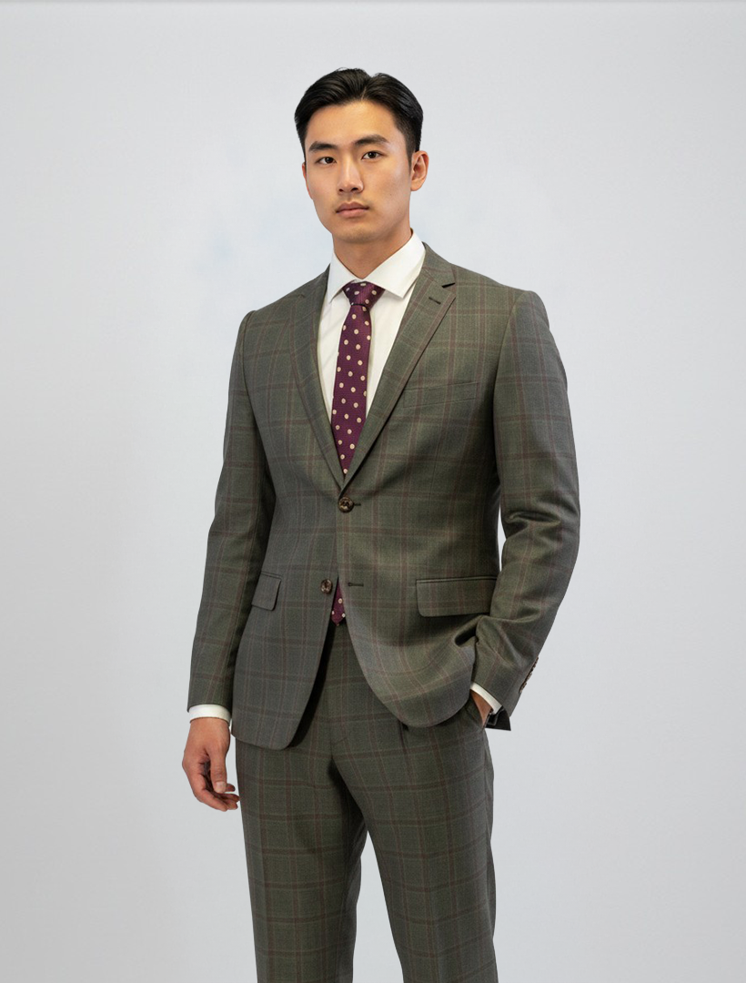 A man wearing the FUBU Olive with Mahogany Windowpane 2-Piece Suit by Fubu The Colle, paired with a white dress shirt and a maroon polka dot tie, stands against a plain light background with a neutral expression.