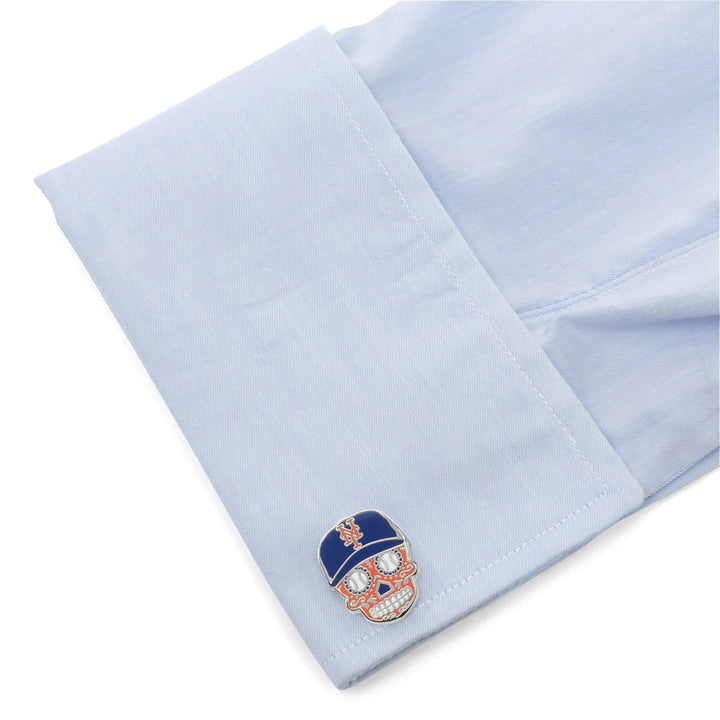 A light blue dress shirt cuff fastened with silver-tone MLB New York Mets Sugar Skull Cufflinks, paired with a blue baseball cap featuring a team logo.