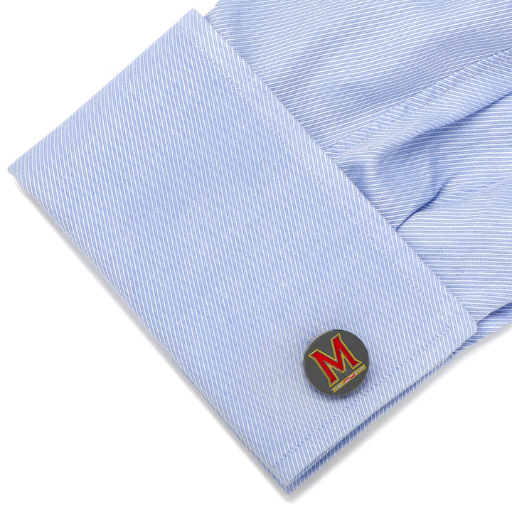 Round silver-toned NCAA University of Maryland Cufflinks with a red letter M logo, fastened to a light blue dress shirt cuff.