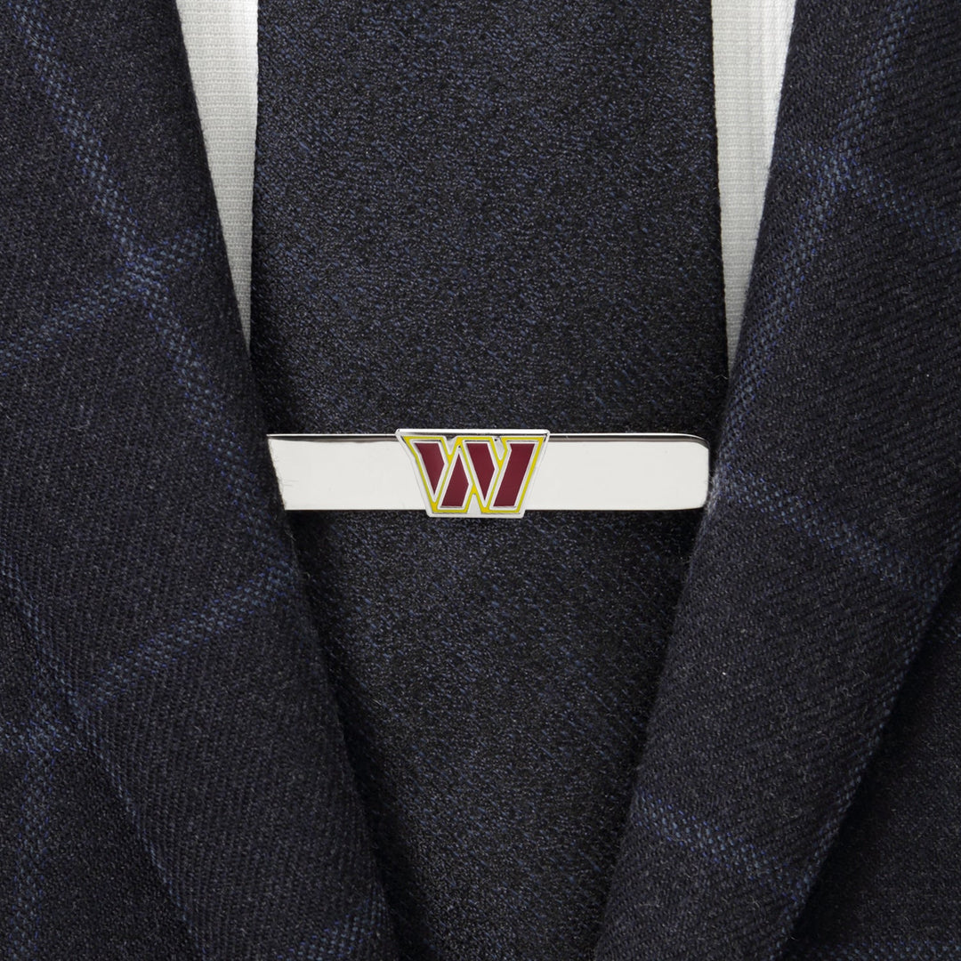 An NFL Washington Commanders Tie Clip featuring a silver “W” logo in burgundy and gold, fastened to a dark blue suit jacket and tie.