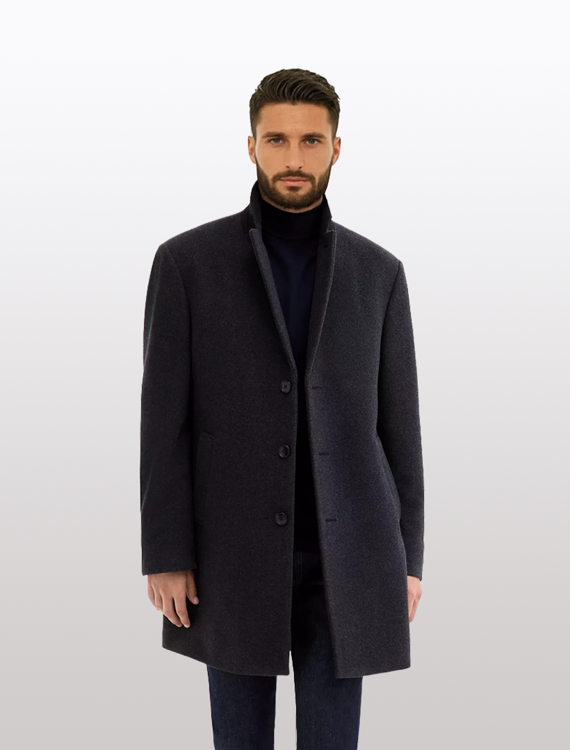 A man models the Calvin Klein Men’s Charcoal Slim-Fit Wool-Blend Overcoat with Notched Lapel over a black turtleneck and dark jeans, standing against a plain light gray background.