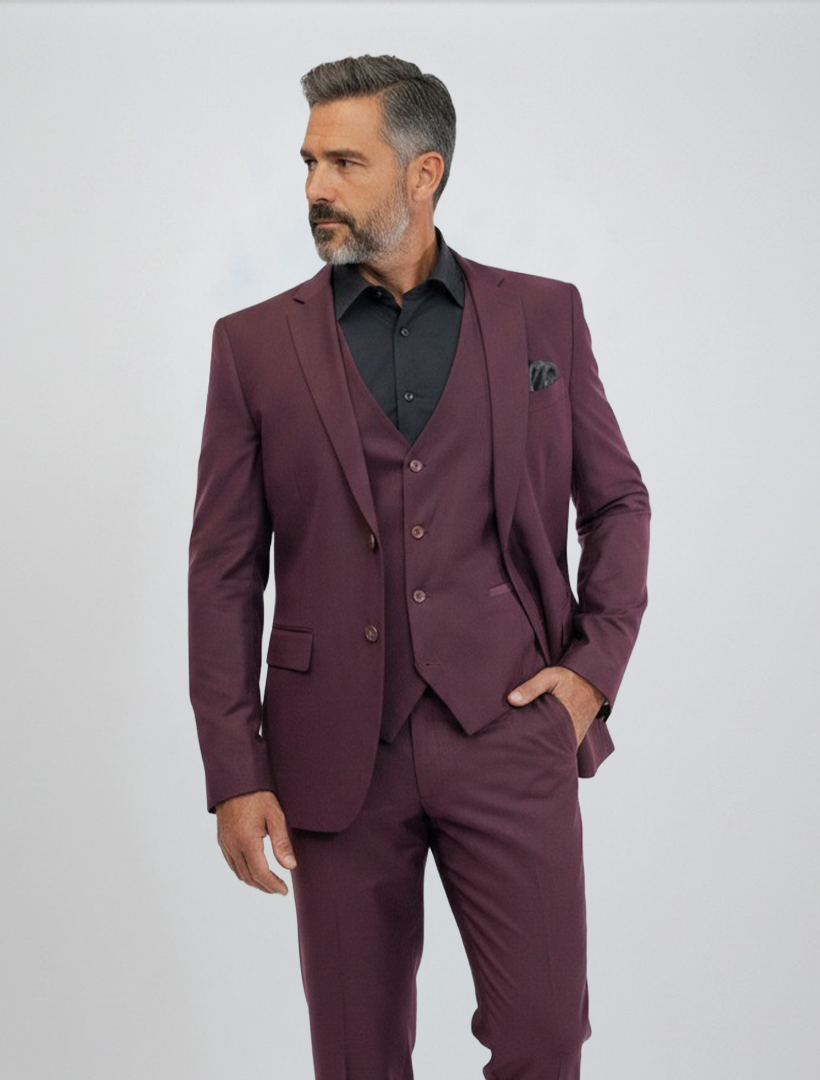 A man wearing the FUBU Burgundy Vested Suit by Fubu The Colle, paired with a black shirt and pocket square, stands against a plain light background looking to his left with one hand in his pocket. This modern fit suit features stretch armholes for added comfort and ease of movement.