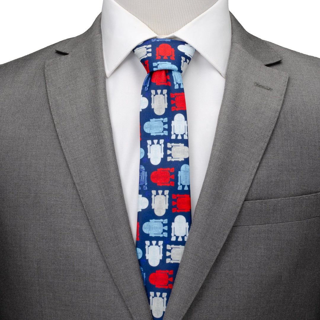 A man wears a gray suit, white dress shirt, and a Star Wars R2-D2 Red, White, and Blue Tie featuring red, white, and light blue droid silhouettes on a dark blue background.