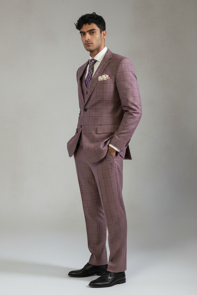 A man wearing the FUBU Mauve Taupe with Purple Windowpane Vested Suit by Fubu The Colle, featuring a slim fit with stretch armhole, is paired with a white shirt, patterned tie, and pocket square. He stands with his hands in his pockets against a light gray background.