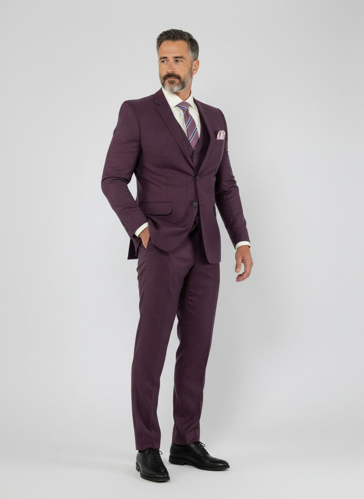 A man wearing the FUBU Purple Solid Vested Suit by Fubu The Colle, paired with a white shirt, patterned tie, and black shoes, stands against a plain light gray background.