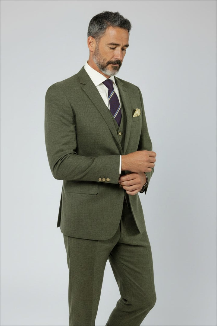 A man with short gray hair and a beard is wearing the FUBU Olive Green Solid Vested Suit by Fubu The Colle, styled with a white shirt, striped tie, and pocket square, standing against a plain light background.