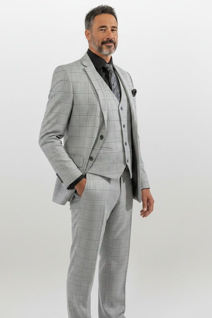 A man wearing the FUBU Light Grey with Charcoal Windowpane Vested Suit from Fubu The Colle, styled with a black shirt and patterned tie, stands against a plain light background. This modern three-piece suit features a slim fit design and stretch armholes for added comfort and style.
