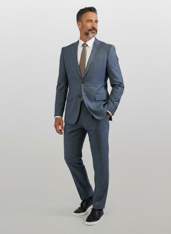 A man stands against a plain light background dressed in the FUBU Gravel Blue with Peach Windowpane 2-Piece Suit from Fubu The Colle, featuring comfortable Tetron Rayon fabric with a stretch armhole, paired with a white shirt, brown tie, and black sneakers.