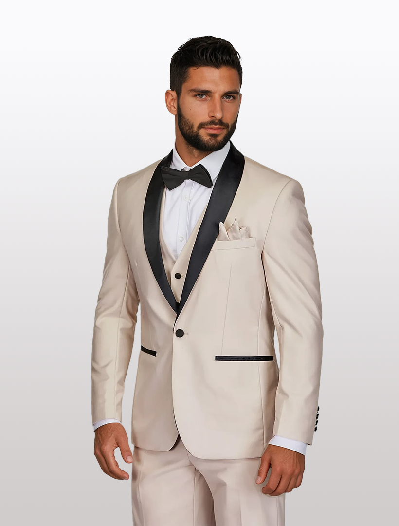 A man in a Gino Vitale Men's Beige Shawl Lapel Tailored Fit Tuxedo with black accents, paired with a white shirt and black bow tie crafted from Tetron Rayon, stands against a light gray background.