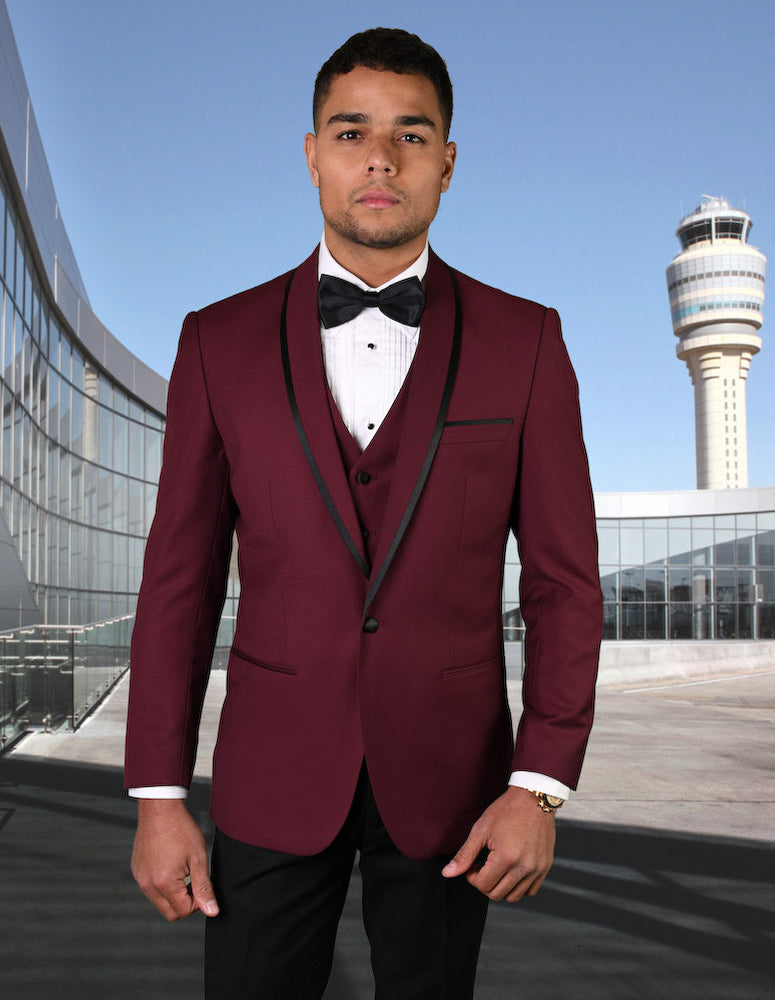 A man in a Statement Men's Burgundy Vested With Black Trim Lapel 100% Wool Tuxedo stands outdoors in front of a glass building and a control tower.